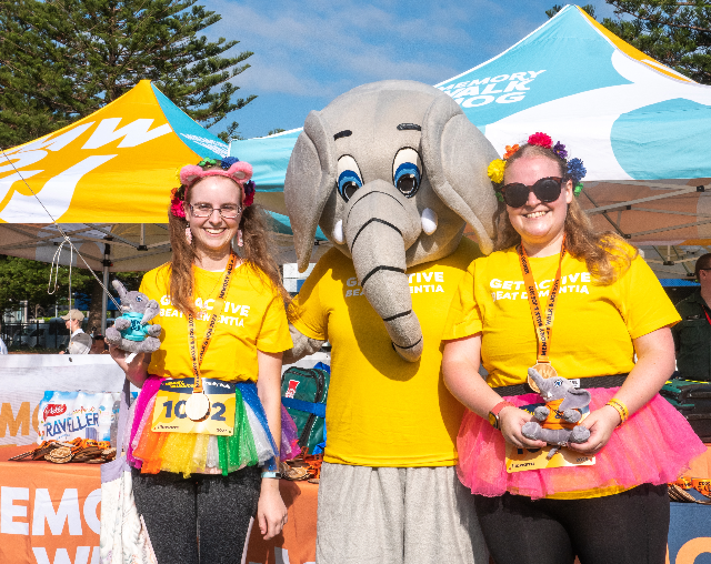 Memory Walk & Jog. Two women participants standing in sun with event mascot, Al the Elephant. Women wearing yellow shirts and colourful tutus.