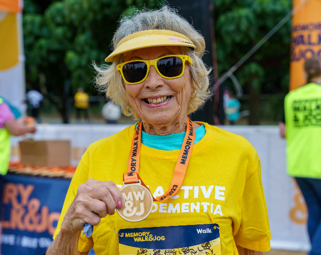 Memory Walk & Jog. Elderly lady smiling and holing medal post-race. Lady is wearing yellow sunglasses, yellow hat, and yellow shirt.