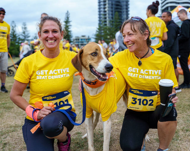 Memory Walk & Jog. Two women kneeling down to have photo taken with dog. Both women smiling and wearing yellow shirts.